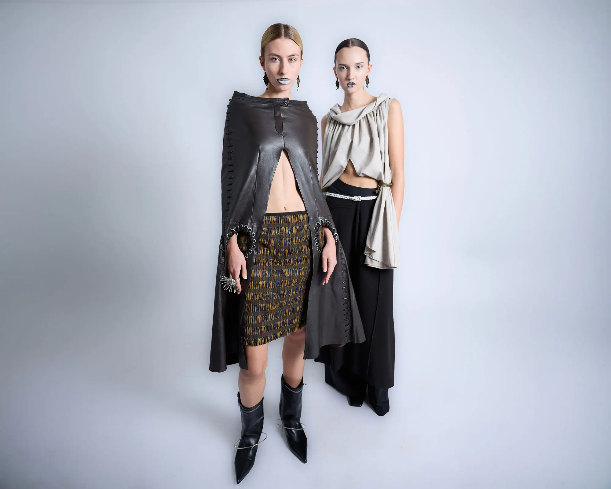 Two models backstage, one in a long black leather cape over a knitted skirt, the other in a grey draped top and black trousers.